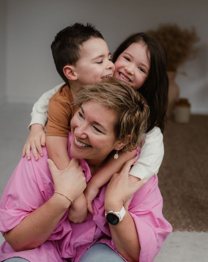 A joyful woman is embraced from behind by two smiling children, creating a warm and playful family moment.